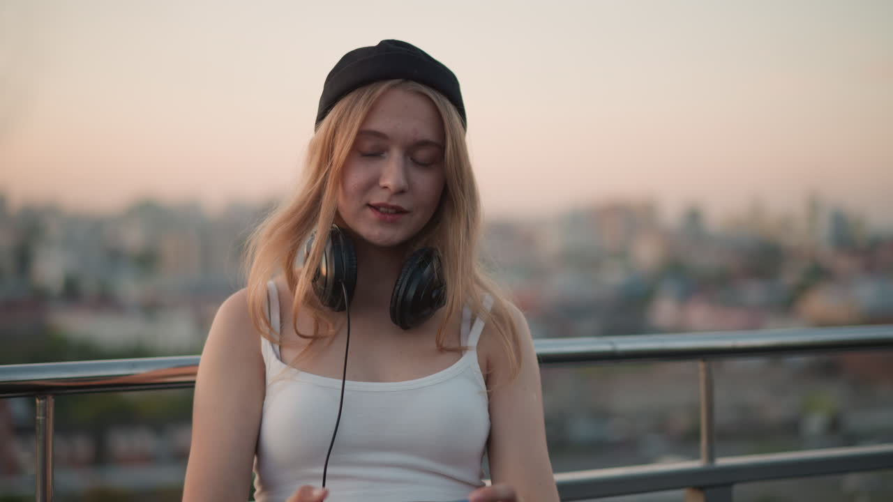 selfie di viaggio al tramonto su un tetto con skyline, giovane donna con cuffie al collo registra un diario di viaggio sul balcone, cielo color pastello, espressione serena, vista panoramica sulla città e luce soffusa della sera