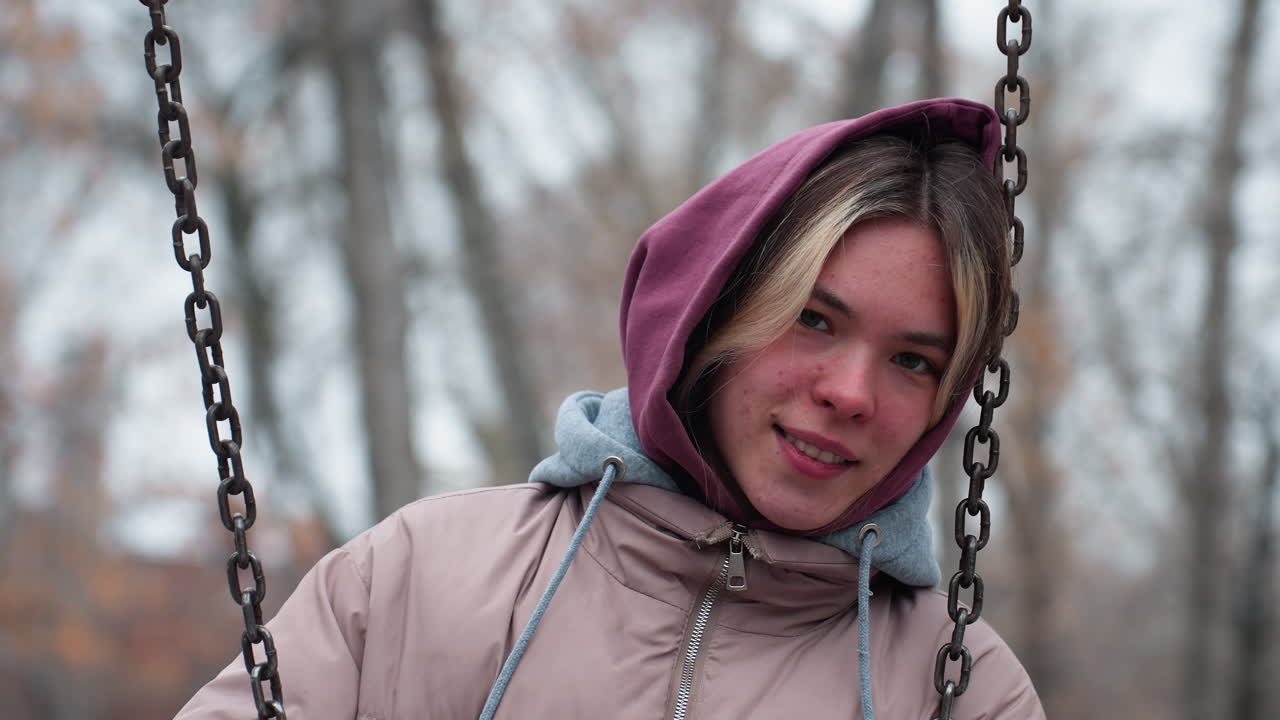 Giovane donna con lentiggini sul viso che indossa abbigliamento invernale, tiene la catena dell'altalena e vi appoggia la testa, vestita con una giacca con cappuccio, guarda dolcemente in lontananza con uno sfondo innevato sfocato.