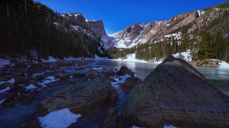 Wide scenic view of a frozen alpine lake surrounded by snow-dusted rocky mountains, evergreen forest, and large foreground boulders under a clear blue winter sky.