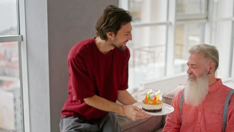 un joven feliz con una morena con paja trae un plato con un pequeño pastel y velas encendidas para su padre anciano con cabello gris y una barba exuberante en una camisa rosa durante sus felicitaciones a su padre en su cumpleaños en un apartamento moderno