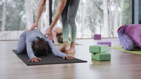 Diverse female instructor assisting woman in practicing child&#039;s yoga pose in studio