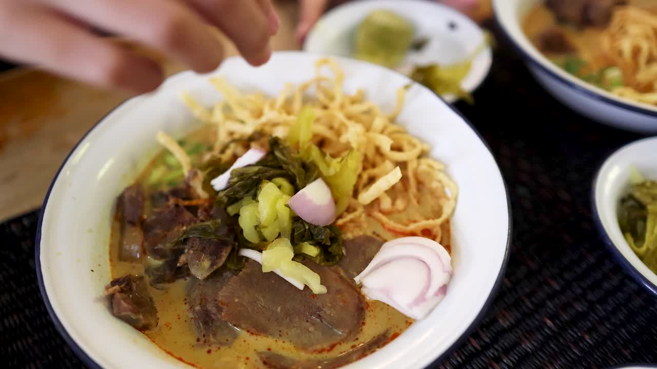 A hand garnishes a bowl of Northern Thai beef curry noodles with sliced shallots and pickled vegetables in a brightly lit restaurant setting, overhead view