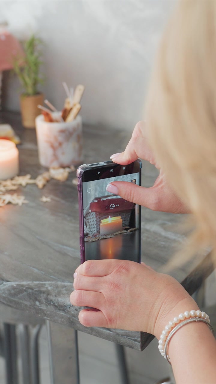 Primo piano di una donna che scatta foto con il suo smartphone, catturando un'immagine con candele e decorazioni rustiche su un tavolo di legno. La scena include una luce soffusa da una candela accesa ed elementi decorativi.