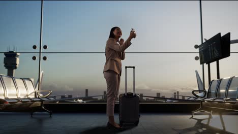 full body of asian businesswoman with rolling suitcase in boarding lounge at the airport, make selfie by smartphone while waiting for flight, airplane takes off outside the window