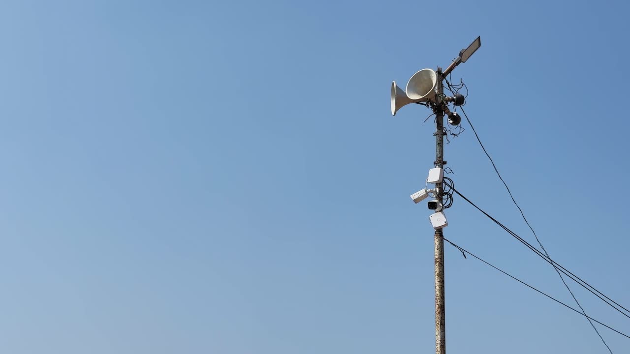 Utility pole with loudspeaker, antenna, solar panel and cables silhouetted against clear blue sky, showing rural communication infrastructure and public address system PA