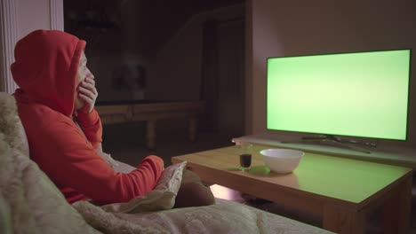 a man in a hood watches the news on tv and switches channels with a remote control, sits on the couch at home