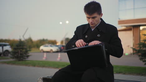 an in black coat seated outdoors after work, going through documents and tablet, holding laminated paper, with background of office building, moving car, street light, and parked car