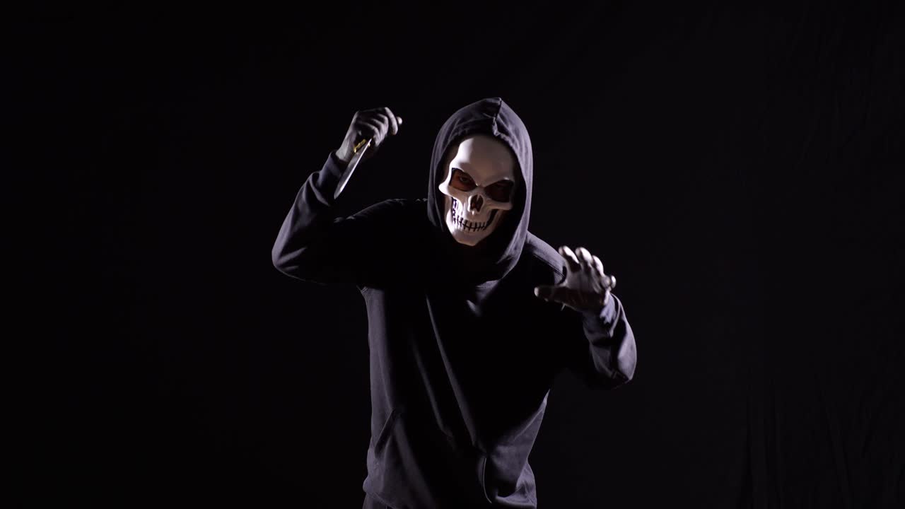 страшный человек в пуловерке с капюшоном, носящий маску хэллоуина, держащий нож и делающий пугающие жесты, бегущий на черном фоне.