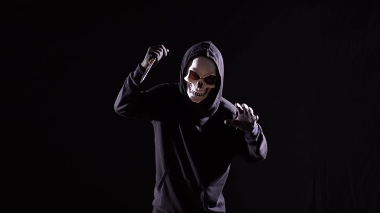 Scary Man In The Hooded Sweatshirt Wearing Halloween Mask Holding A Knife And Making Frightening Gesture While Running On The Black Background