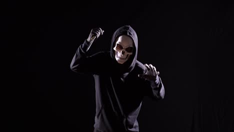 hombre aterrador en la sudadera con capucha usando una máscara de halloween sosteniendo un cuchillo y haciendo un gesto aterrador mientras corre sobre el fondo negro