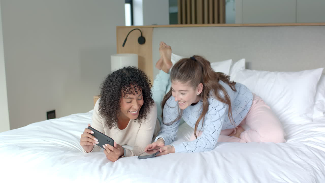 Using smartphones, two women relaxing on bed and smiling at home