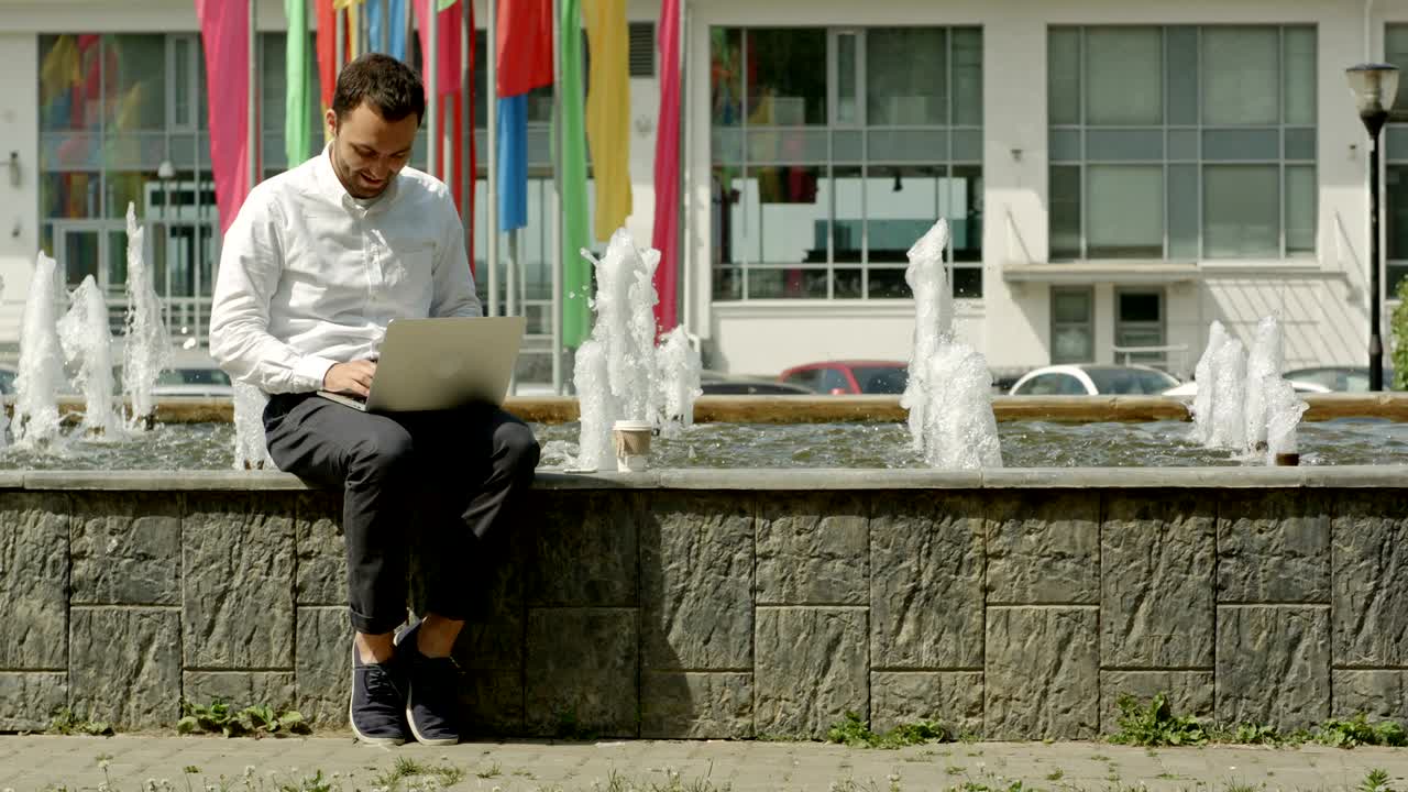 homem com laptop sentado perto da fonte e feliz com os resultados de seu trabalho