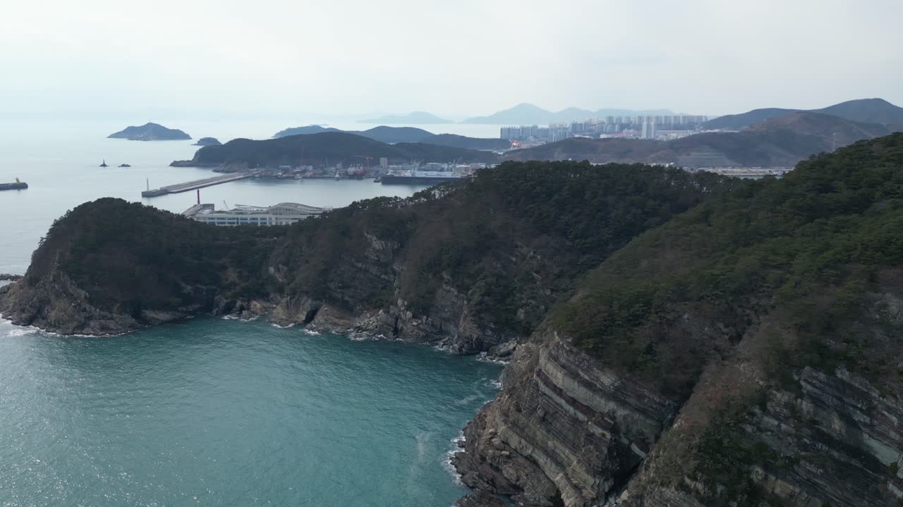 Drone aerial view in South Korea countryside flying over the clear blue sea of Busan next to a green mountain, ships and city in the background sunny day