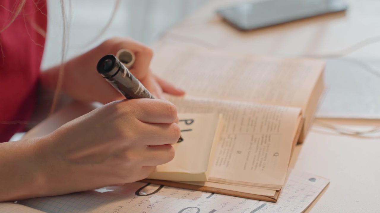 開いた本に挟まれた付箋に書き込む若い女性。手書きのメモが散らばった書類が写る。柔らかな自然光が、様々な学習用品が置かれたこの机のセットアップを照らしている。