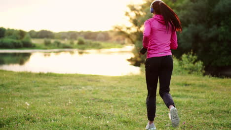 brunette with long hair in headphones runs along the river in the park in the morning at sunrise in the summer in a pink jacket and black pants