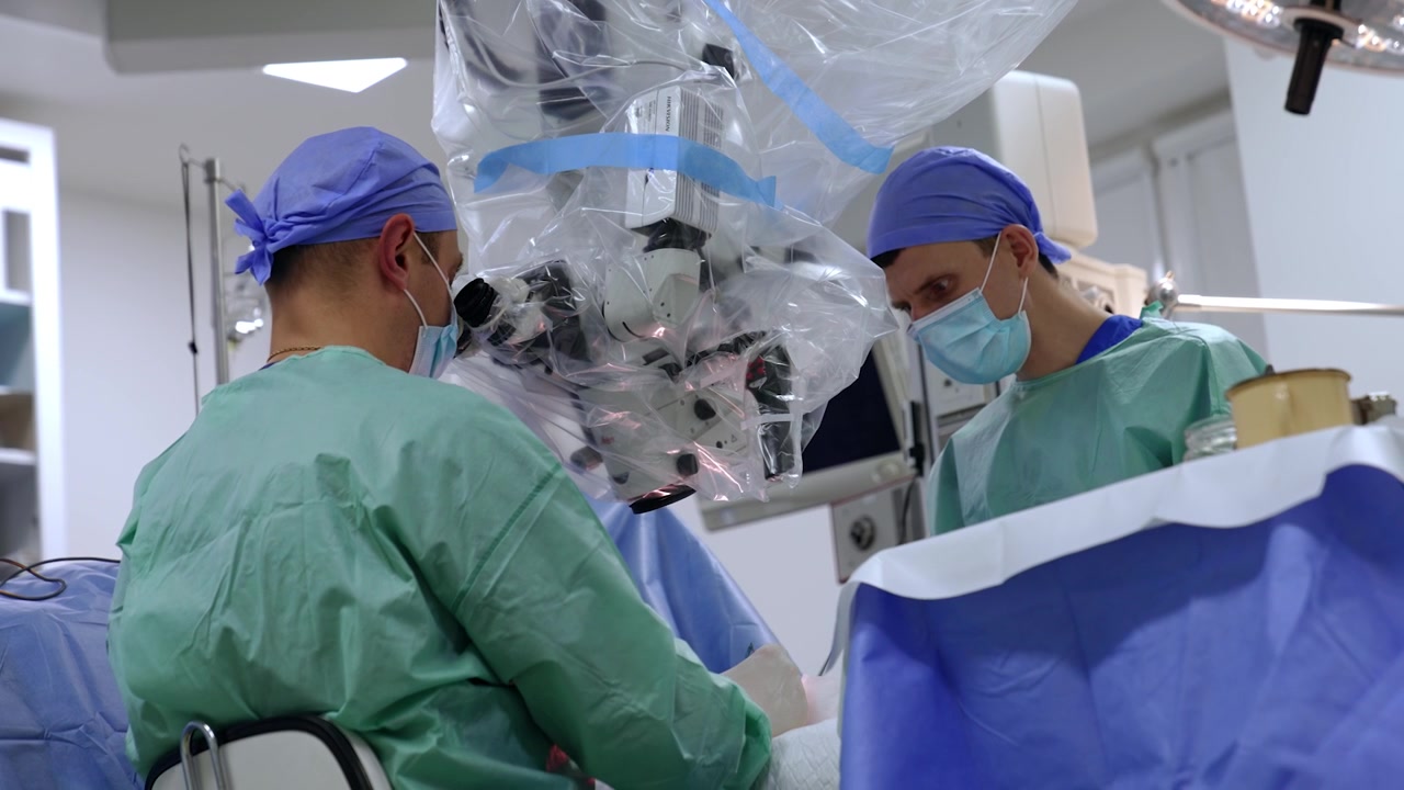 Mid-aged male surgeon sits at the microscope carrying out microsurgery. Rear view. Doctor passes the used tool to the assistant standing opposite. Low angle view.