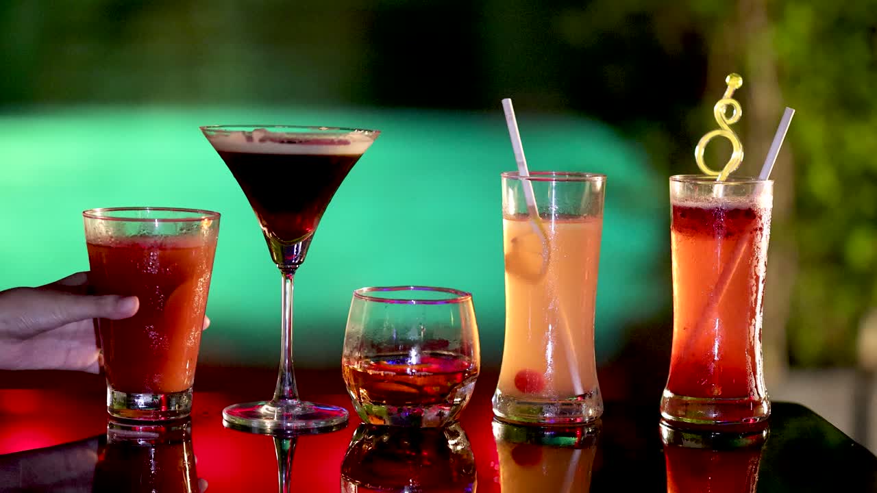 Multiple hands choose cocktails from a vibrant bar table, illuminated by dramatic colored lighting
