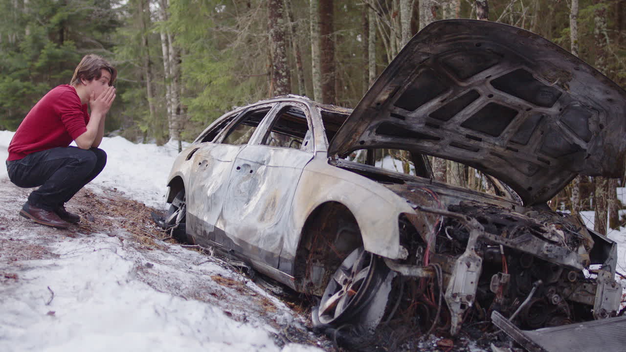 Devastated student with red jersey squats near wrecked car next to icy road