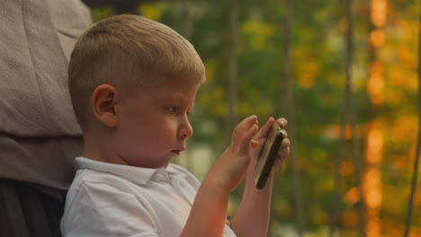 blond boy enjoys pastime with smartphone on balcony. adorable child focuses on interesting game sitting in armchair with breathtaking view of countryside