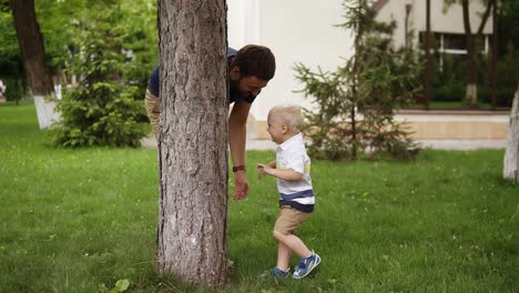 niño pequeño divirtiéndose con su padre en un parque verde. jugando a buscar y esconderse. niño lindo y divertido escondiéndose detrás del árbol y el padre lo atrapó. tiempo de verano. cámara lenta