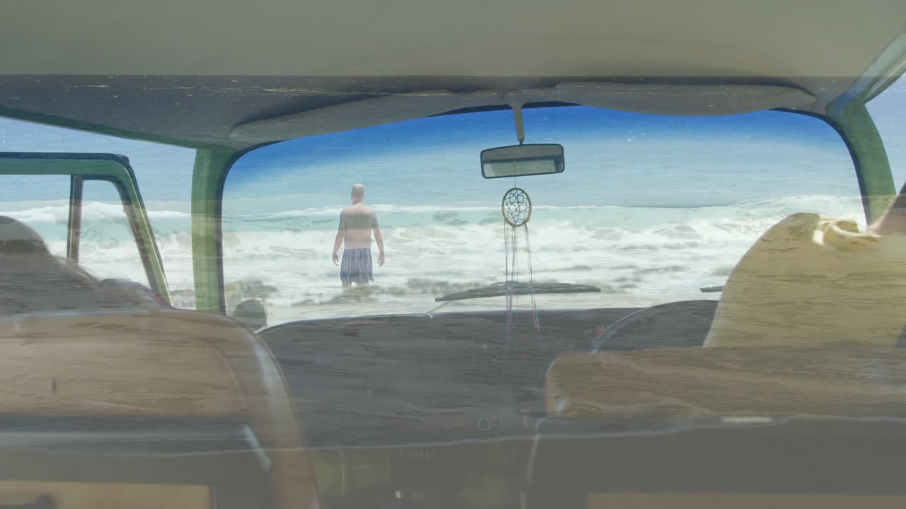 Walking into ocean, man seen through vintage car windshield with dreamcatcher hanging