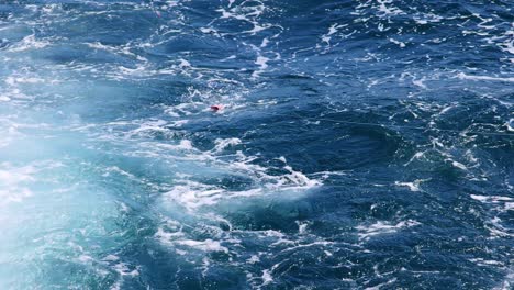 a red object floats amidst turbulent ocean waves, captured from a moving boat under bright daylight