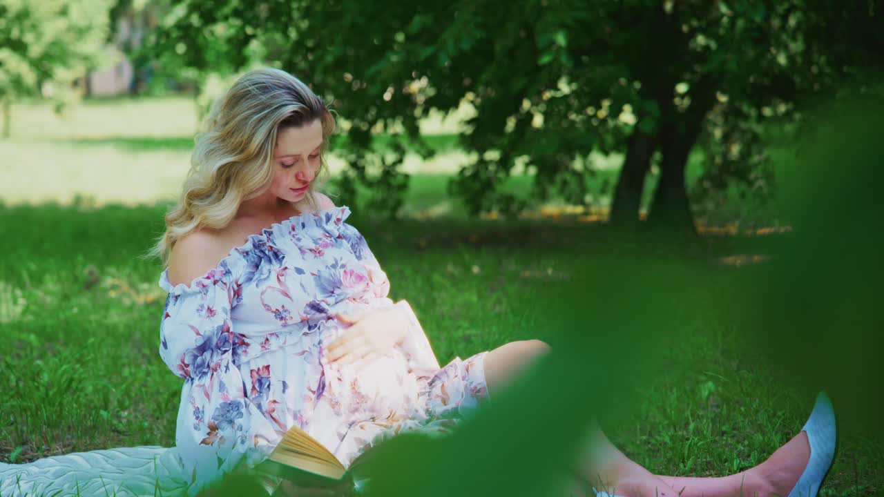 Gravid kvinde læser en bog og rører ved sin mave i en park