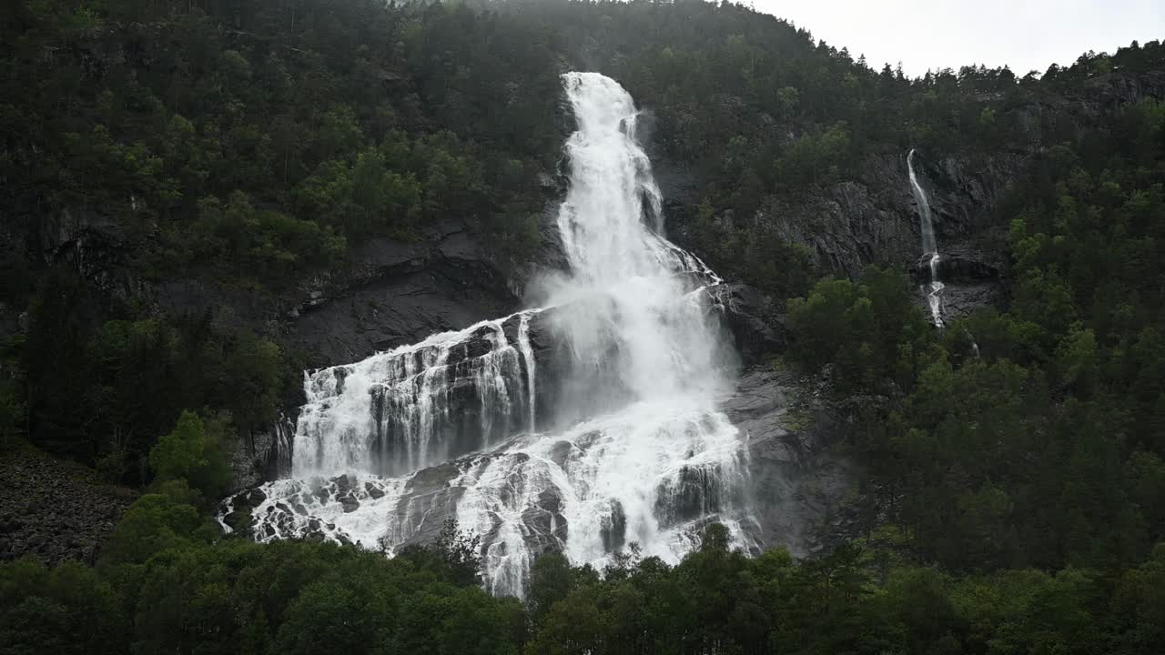 Slow motion of powerful waterfall surging down green mountainside in Voss during heavy rain and flooding. Lush summer landscape under stormy conditions