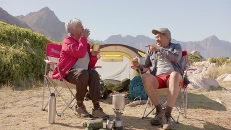 happy senior biracial couple sitting at tent in mountains and drinking coffee, in slow motion