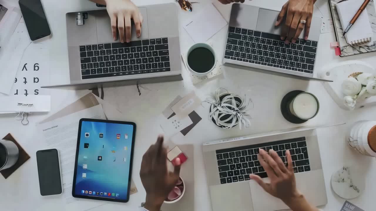 Top-down video shot of diverse hands working on laptops and tablets at a cluttered desk