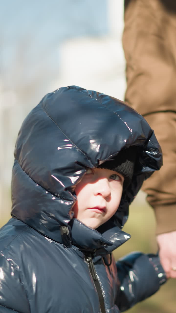 padre e hijo caminando de la mano al aire libre, el niño, vestido con una chaqueta negra brillante, mira hacia arriba mientras caminan juntos, con un fondo borroso de árboles