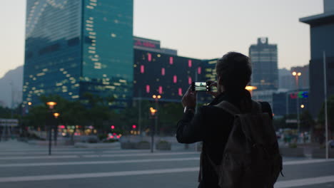 Junger-Kaukasischer-Touristenmann,-Der-Mit-Dem-Smartphone-Ein-Foto-Des-Wunderschönen-Bürogebäudes-Mit-Der-Skyline-Der-Stadt-Macht-Und-Abendliche-Stadtreisen-Genießt