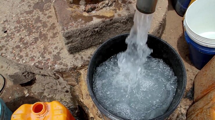 Un primo piano di acqua limpida che sgorga da un tubo di metallo in un secchio di plastica nero, con altri contenitori visibili nelle vicinanze a terra.