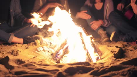 Closeup-view-of-the-bonfire-late-at-night.-Young-people-sitting-by-the-fire-in-the-evening,-playing-guitar.-Cheerful-friends-singing-songs,-talking-and-having-fun-together