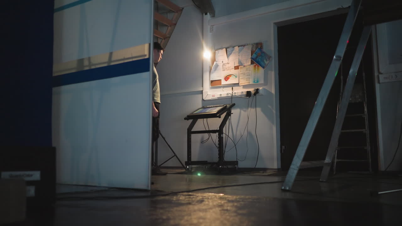 Man works on illuminated touchscreen console in dimly lit technical room under stairs, surrounded by wires, tools, light source, ladders, and detailed wall notes, showing quiet focus in backstage setup