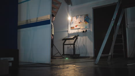 man works on illuminated touchscreen console in dimly lit technical room under stairs, surrounded by wires, tools, light source, ladders, and detailed wall notes, showing quiet focus in backstage setup