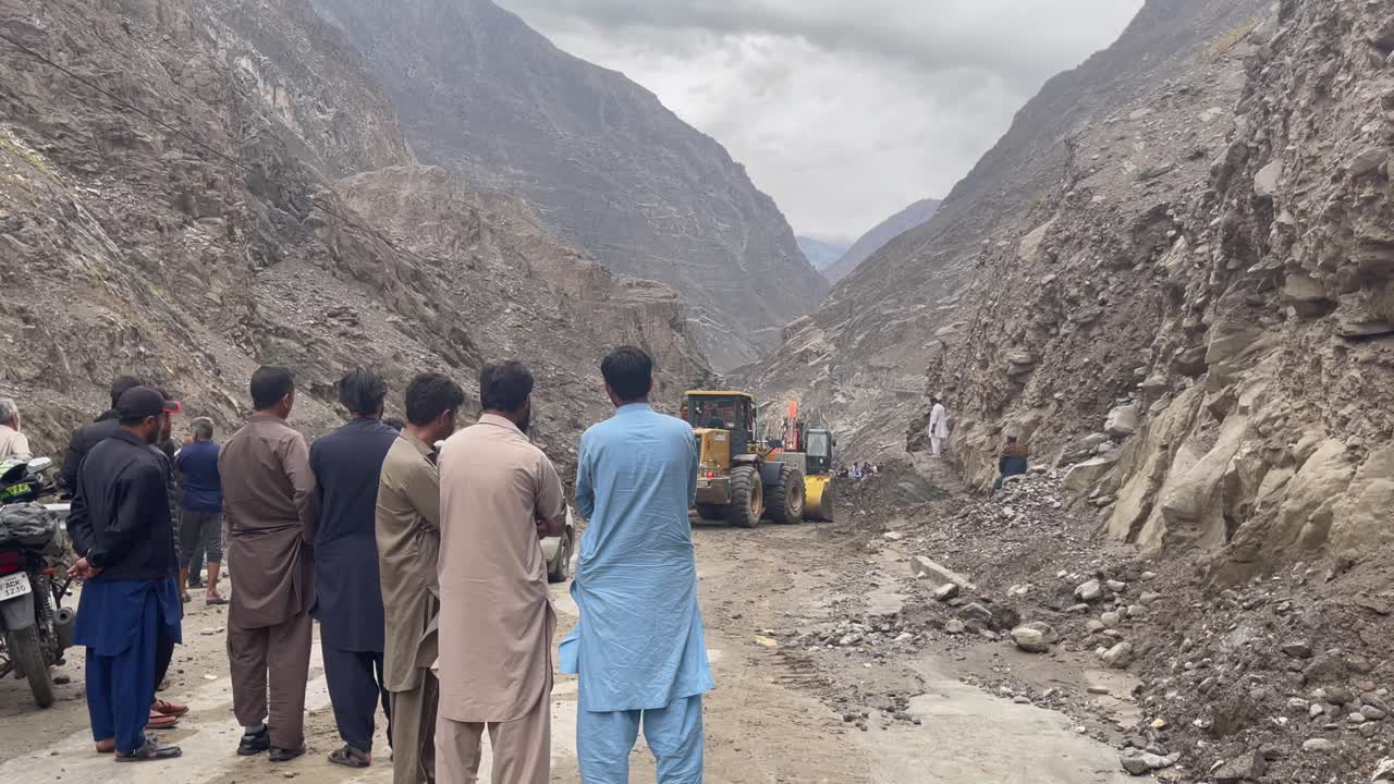 A group of locals watch as a bulldozer works to clear a landslide blocking the Gilgit Skardu Road in Pakistan. The scene depicts community efforts and perseverance in overcoming natural obstacles.