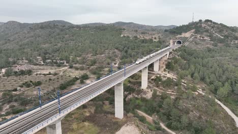 a bullet train is exiting a tunnel and moving along the concrete bridge in the middle of a beautiful landscape in rural spain