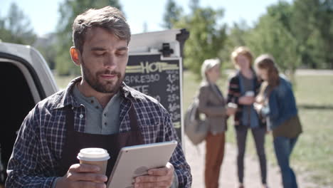 koffie vrachtwagenchauffeur terwijl hij een tablet gebruikt en koffie drinkt, een groep mensen praat en drinkt koffie op de achtergrond