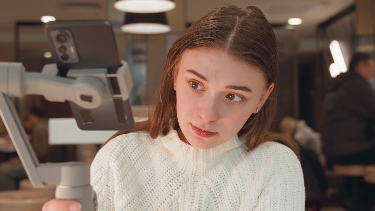 Mid-close scene shows young woman holding smartphone on handheld gimbal, subtly shifting wrist and turning head with thoughtful focus, ready for mobile recording inside cozy cafe setting
