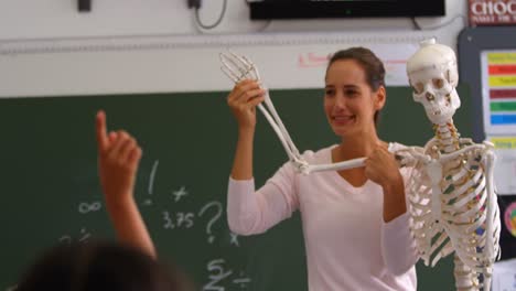 vista frontal de una maestra caucásica explicando el modelo de esqueleto humano en el aula 4k