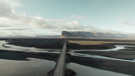 flyover above bridge on highway running through black sand desert in iceland towards rock formation