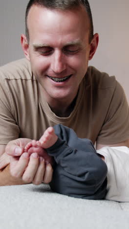 smiling caucasian man sits near his sleeping infant son. father holds and strokes tiny feet of his baby and kisses them. vertical video