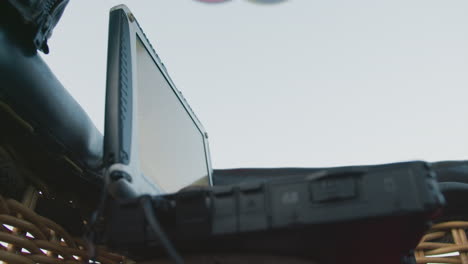 close up of rugged navigation laptop with open screen showing faint map lines placed inside hot air balloon basket under sky suggesting travel monitoring and flight direction