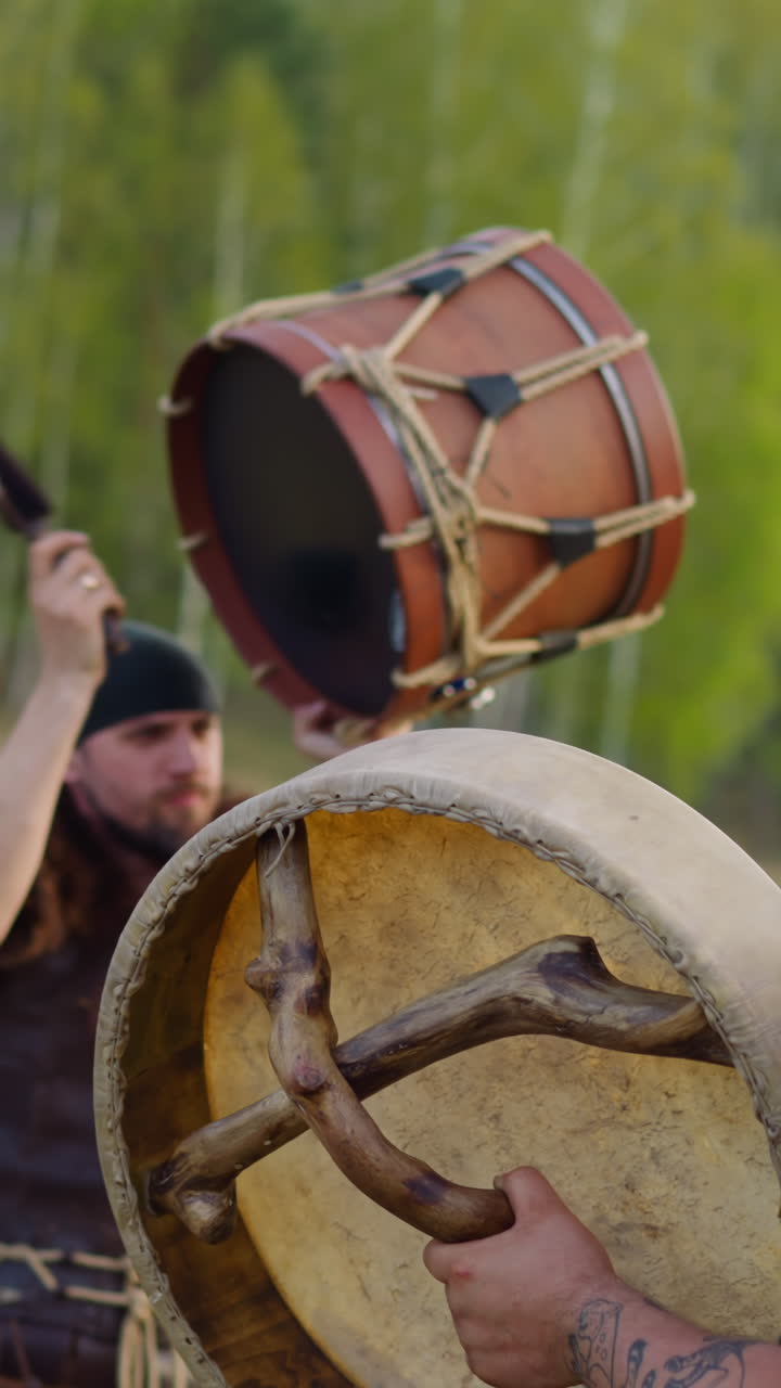 Man in folk clothes taps drum with friend against green forest in spring. Ancient spiritual ceremony at wild nature. Sound for meditative practices