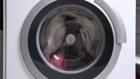 closed drum of the washing machine rotates slowly rinsing dirty multi-colored laundry with fabric softener, close up. advertising of large household appliances for general cleaning of the house