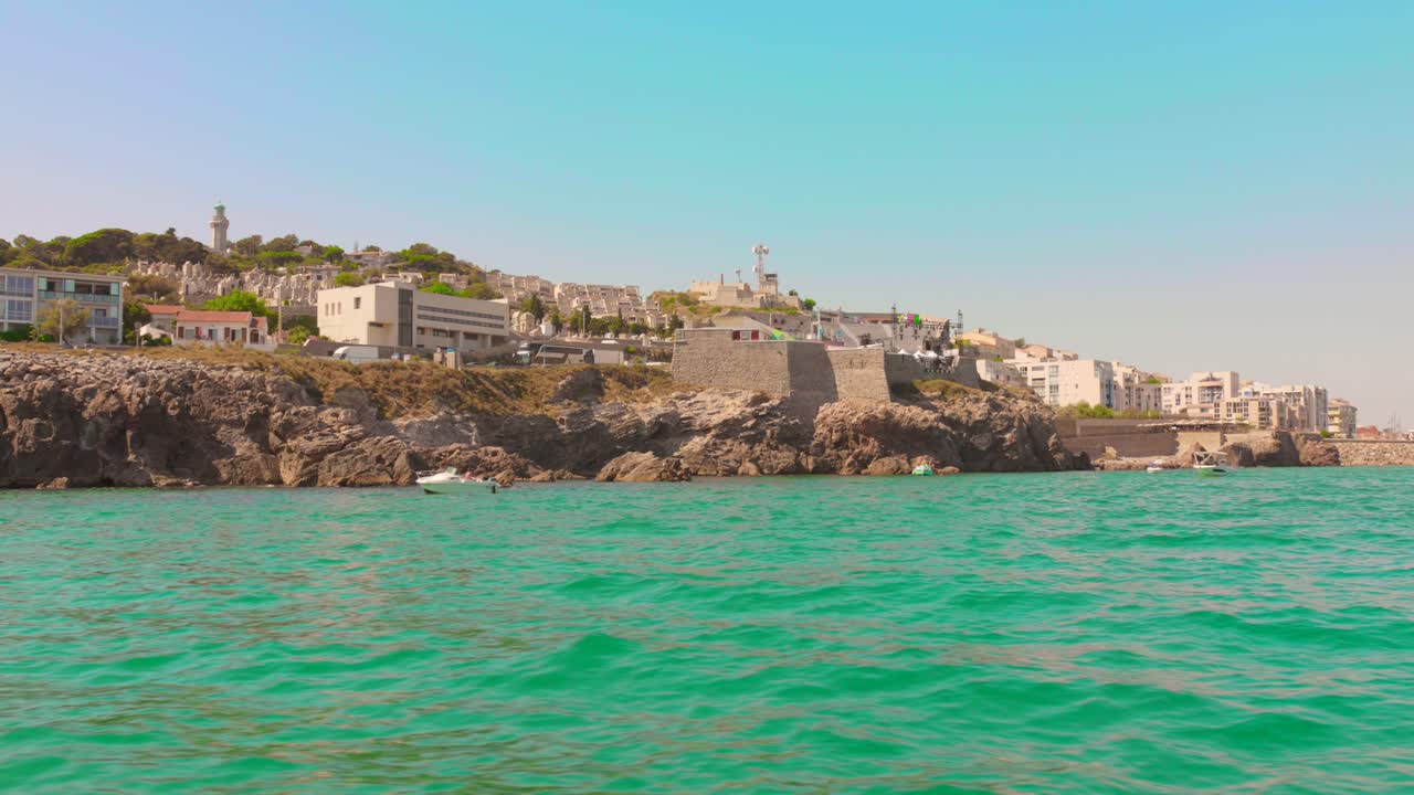 Pan left of Sète's rocky coast featuring historic fortifications and hillside structures seen from the water on a clear summer day