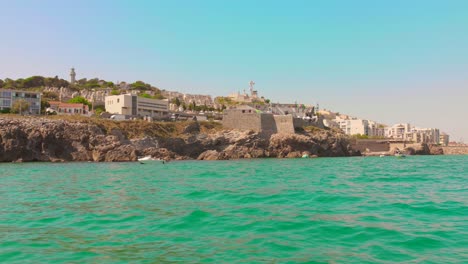 pan left of sète's rocky coast featuring historic fortifications and hillside structures seen from the water on a clear summer day