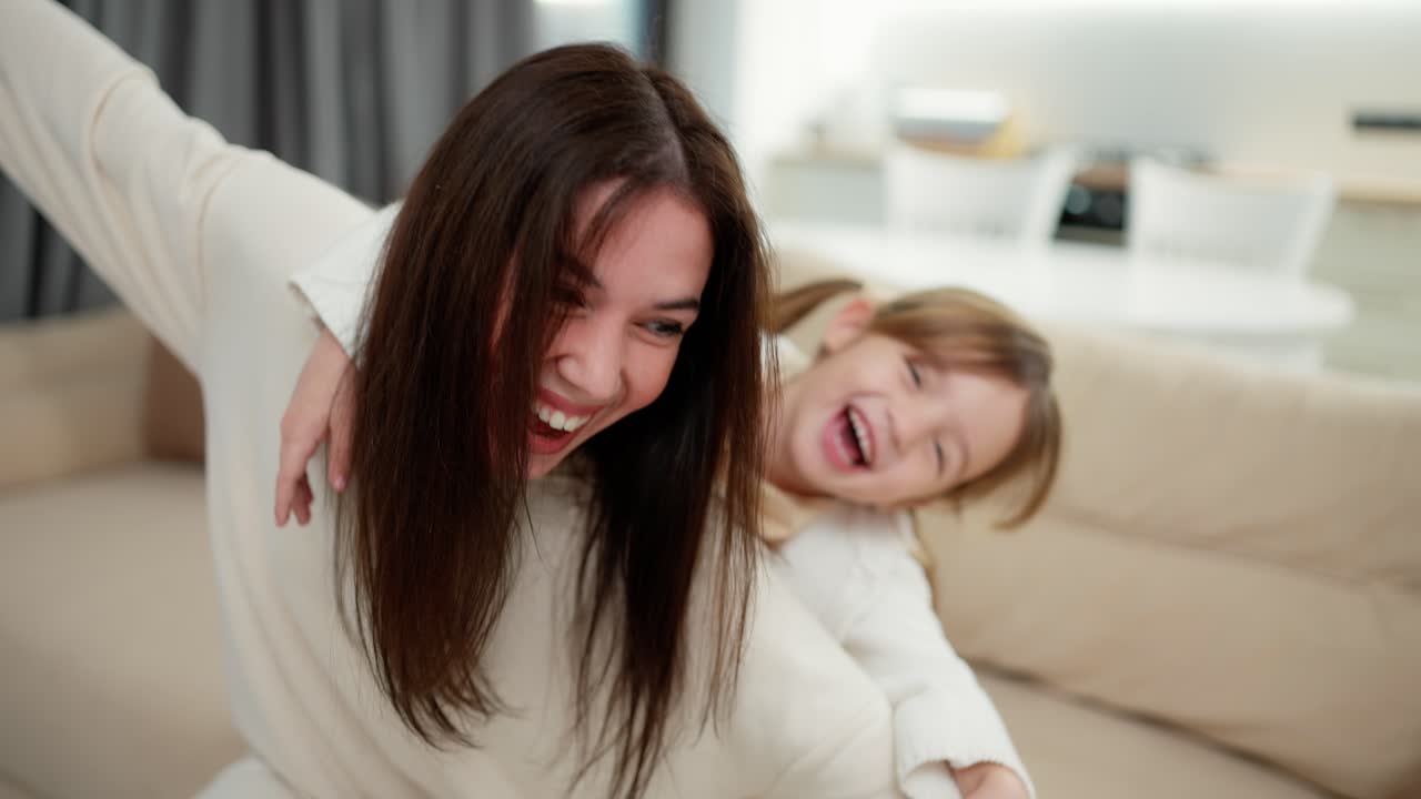 즐거운 어머니가 작은 아이 소녀를 등에  ⁇ 고 함께 즐겁게 날아가는 척하고 있습니다.