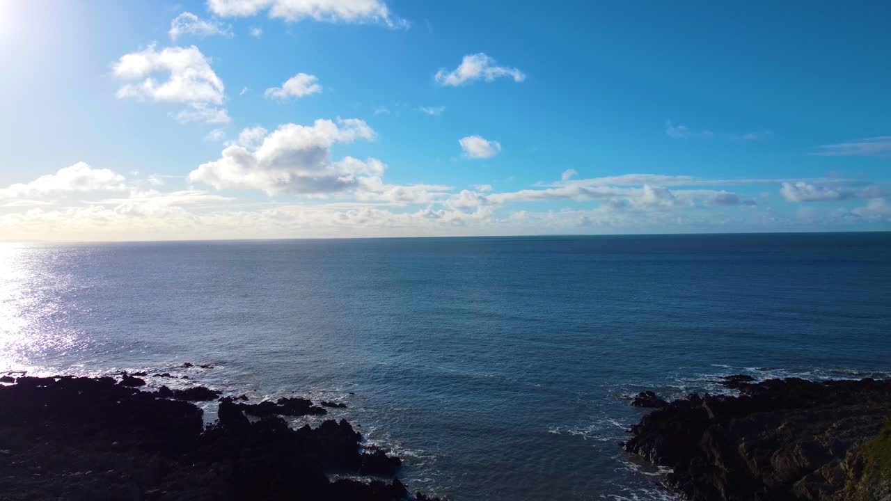 Lenta carrellata da una spiaggia rocciosa che si dirige verso un tranquillo paesaggio marino con il sole che si riflette sulla superficie dell'oceano. Drone della costa di Gower nel Galles del Sud, Regno Unito. Perfetto per video di meditazione rilassanti